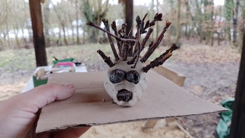 a clay deer with twig antlers held on a piece of cardboard