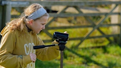 A young girl wearing a yellow sweatshirt looking sideways on at a handheld camera on a tripod