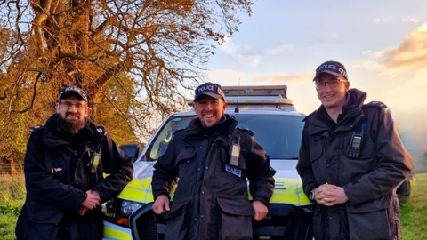 Three members of Norfolk Police standing smiling in front of a police car beneath a tree in autumn colours