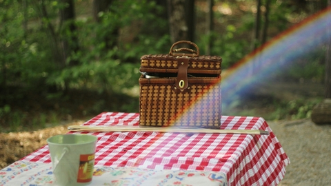 A wicker picnic basket standing on a red and white checked tablecloth. A rainbow is across photo