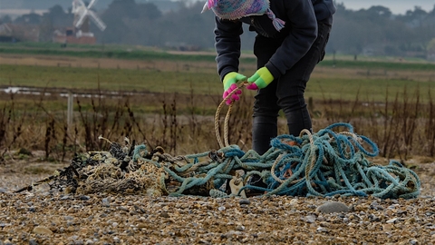 A lady wearing green gloves and a woolly hat bending over and removing rope lying on the beach