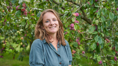 Kate Bradbury standing smiling under a plum tree and wearing a blue dress