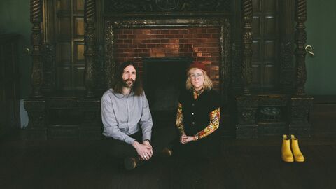 Musicians Samantha Whates and MG Boulter are seated in front of a large open fireplace in a dark, historic room