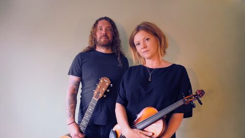 Georgia and Aaren standing together wearing blue t-shirts and holding a guitar and violin