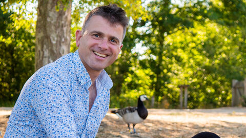 Nick Acheson is wearing a blue and white shirt and blue shorts, smiling at the camera as he crouches down to feed a Canada goose