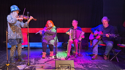 Stookey Blue ceilidh band members in a dark venue playing a fiddle, guitar, flute and accordionn