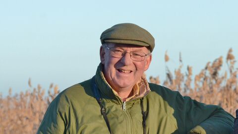 Bernard is leaning on a wooden gate, wearing a green coat and cap and has a pair of binoculars around his neck