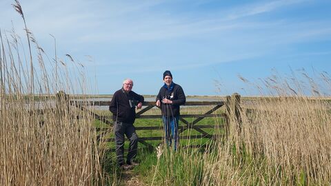Two men stand in front of a gate in the middle of Cley Marshes. They are both looking at the camera and smiling. They are dressed in black fleeces and combat trousers, with binoculars around their necks.