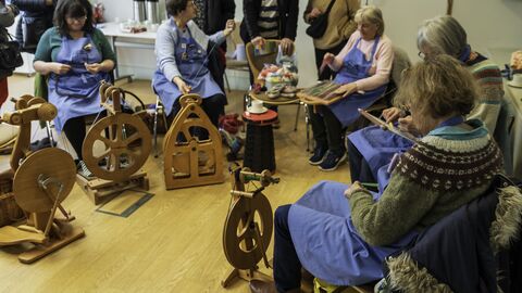 A group of spinners wearing blue aprons sitting in a semi-circle spinning yarn on wooden looms