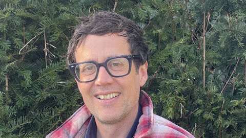 Patrick Barkham is wearing a red and white checked shirt, dark-rimmed glasses and is smiling at the camera
