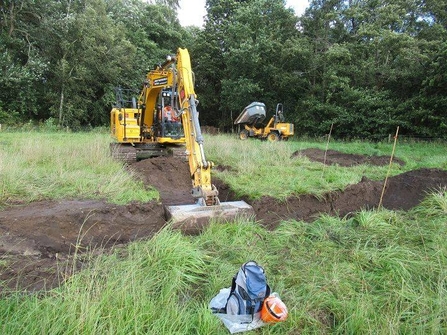 A digger works on excavating a ghost pingo at Watering Farm. The digger digs earth out of a long ditch, while a tip truck sits in the background.