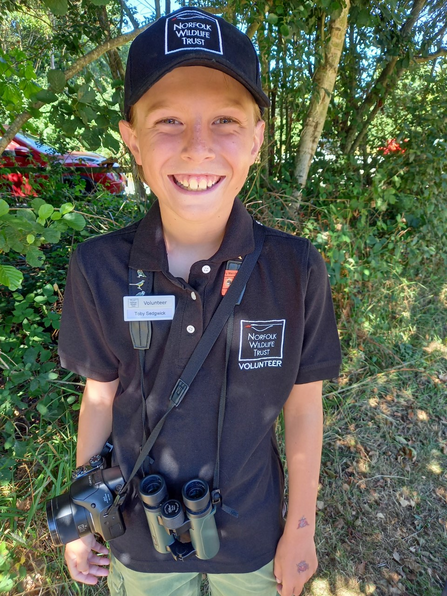 A young boy smiling and wearing his NWT t-shirt, cap and name badge. He is also carrying a pair of binoculars and a camera.