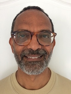 A volunteer smiling at the camera. He has a grey stubbly beard, black hair and round tortoiseshell glasses.