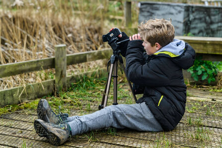A blond-haired boy wearing a black coat and grey trousers sits on the ground looking through a camera on a tripod