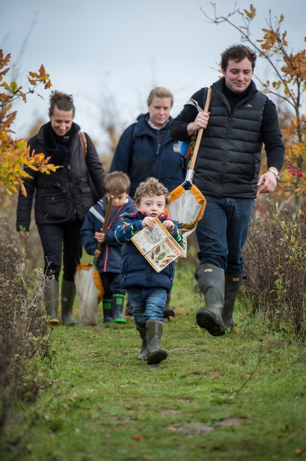 A family carrying ID guides and nets walking to a pond to do some pond dipping. They are wearing warm coats and welly boots.
