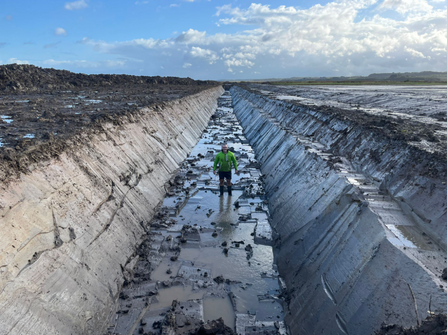 Reserve staff member, Adam, standing with his arms outstretched in the new, New Cut that had just been dug at Cley. It's a huge channel cut through the earth, Adam looks tiny standing in it.
