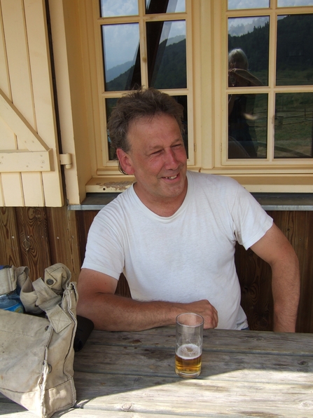 John Allaway smiling on a wooden bench with a beer