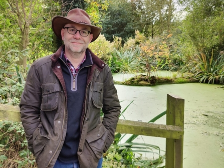 Richard is wearing a big brown hat and is smiling in front of a green body of water with lots of trees and shrubs around.
