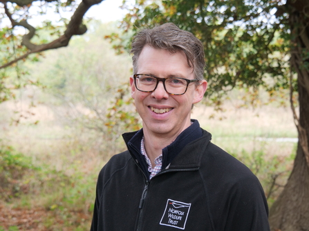 Norfolk Wildlife Trust CEO, Eliot Lyne, smiles at the camera while standing in front of woodland. He is wearing glasses and a black NWT fleece jacket