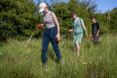 Three young people with gloves, bin bags and pickers, looking for rubbish at Soughbottom Meadows on a sunny day.
