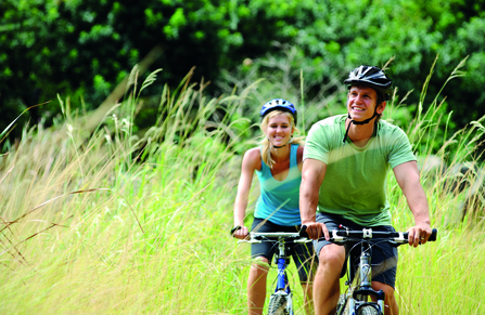 A couple smiling and cycling on a sunny day surrounded by tall grass. 