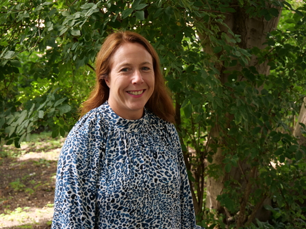 A woman with reddish hair smiling in a park, surrounded by trees. She is wearing a blue polka dot top. 