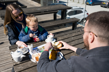 A family with a toddler enjoying cake and tea on benches outside the visitor centre at Cley in the sunshine.
