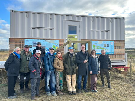 A group of smiley people in warm coats and hats, some carrying binoculars, stood outside the new Gilmor Discovery Hide at Cley