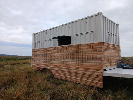 A close up of a wildlife hide on wheels. It is a metal container with wood panelling on the bottom half.