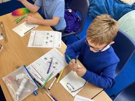 A young boy in class, he is wearing glasses and uniform and is filling in sheets with coloured pens. 