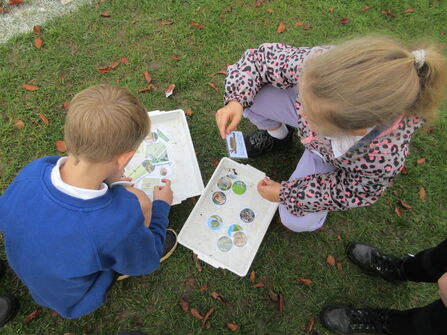 Two primary school children crouched on the ground looking through activity sheets and photos of different habitats.