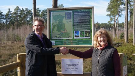 A man and woman, smiling and shaking hands in front of Broadland Country Park reserve signage.