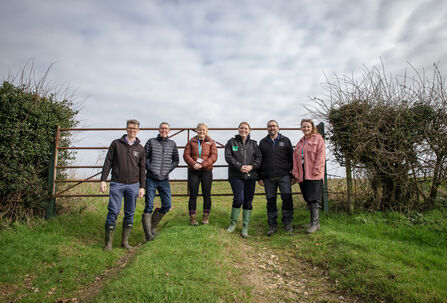 A group of people in wellies smiling and posing together in front of a farm gate. 