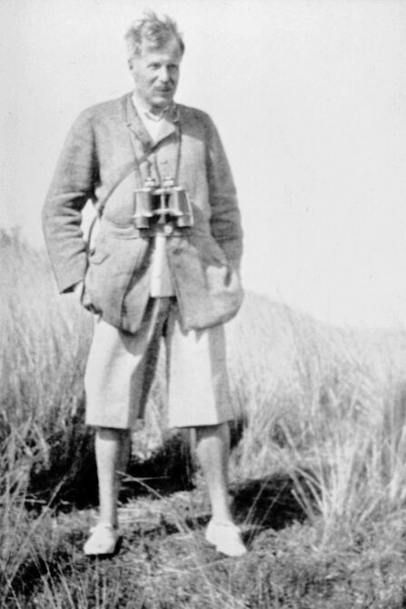 An old black and white image of a man in a linen suit and shorts with a pair of binoculars. 