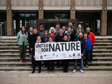A group of smiling NWT staff on the steps outside Nofolk County Hall holding up a banner that says 'united for nature'.