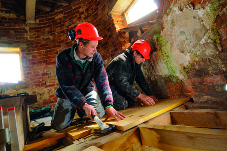 Two men in hardhars putting down wooden flooring in an old stone building. 