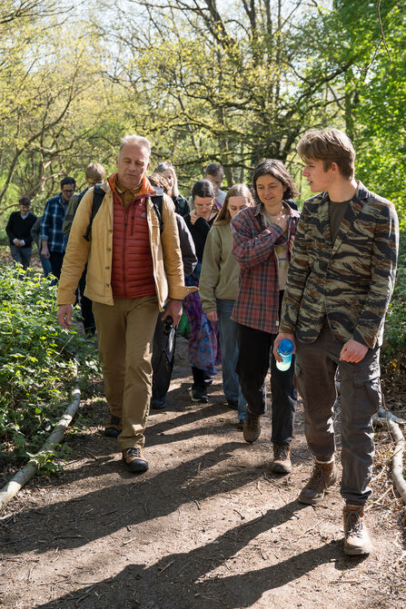 A man wearing a yellow coat and red puffa jacket leads a line of young people in a walk through woodland on a sunny day