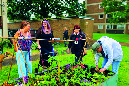 A group of people tending to a community garden, on a piece of grass beside some blocks of flats. One man wearing a hat is leaning down and tending to some flowers, while three women wearing bright clothing look on. One of them is wearing a flower crown and has a mobility aid.