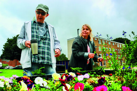 Two people garden in a raised bed full of flowers, with a tall block of flats visible in the background, under a grey sky. The person on the left is wearing dark glasses and holding a thermos, while the person on the right is holding a leaf.