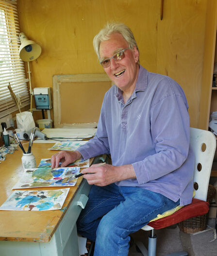 Artist John Hurst smiles at the camera as he paints at his desk