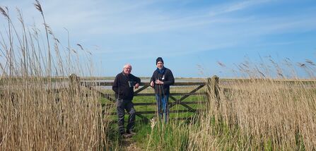 Two men stand in front of a gate in the middle of Cley Marshes. They are both looking at the camera and smiling. They are dressed in black fleeces and combat trousers, with binoculars around their necks.