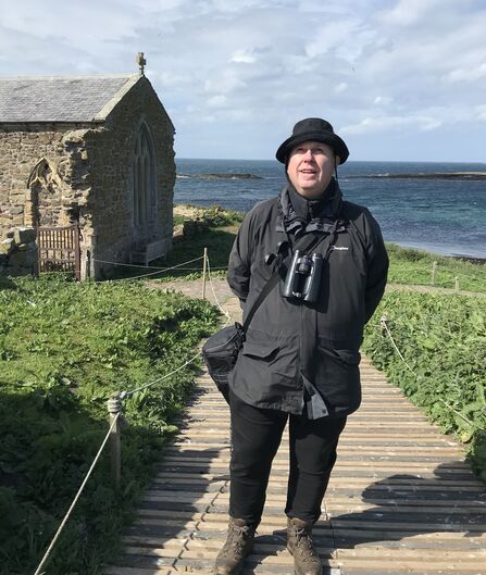 A man all in black with binoculars around his neck is smiling on a boardwalk beside the sea. There is a small chapel in the background.