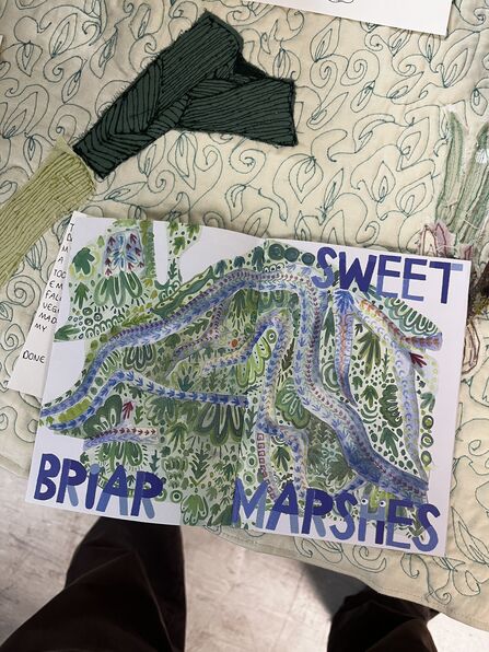A hand drawn map of Sweet Briar Marshes, in green and blue colours, resting on a table covered in a cream and green tablecloth.