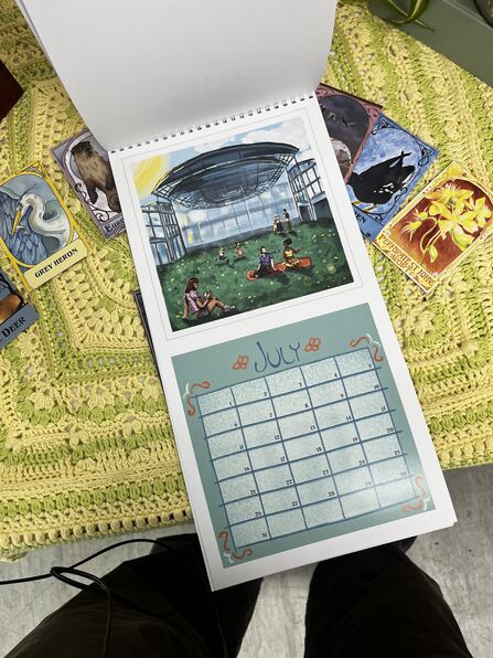 A hand drawn calendar lying open on a table covered in a yellow and green blanket. The calendar is open to the July page. There are also cards featuring animals around the calendar.