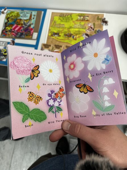 A hand holding a bright and colourful booklet, opened up to a page with a pink and purple background and featuring plant and butterfly drawings.