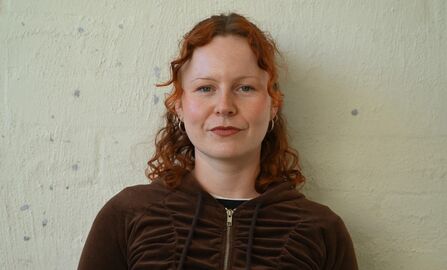 A head and shoulders photo of a young woman smiling at the camera. She has curly, red, shoulder-length hair and is wearing a brown zip-up jacket. She stands in front of a brick wall, which is painted white.