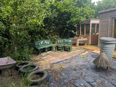 Benches with bunting, old tires, a wheelbarrow, compost bin and trees on school ground. 
