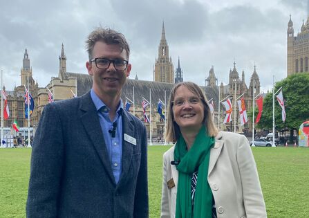 A man and a woman in suits, smiling outside parliament.