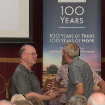 Two men shake hands in front of a roller banner reading '100 years of trust, 100 years of hope'. One of the men is holding a small wooden trophy.