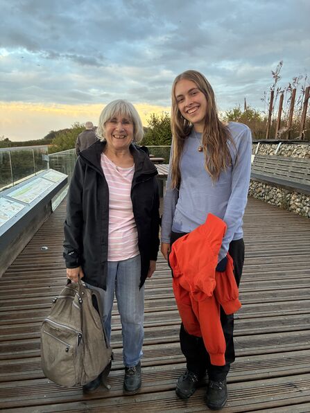 Two volunteers stand side by side, smiling, on a viewing platform in front of the sunset. They are both wearing coats, jumpers and walking boots.